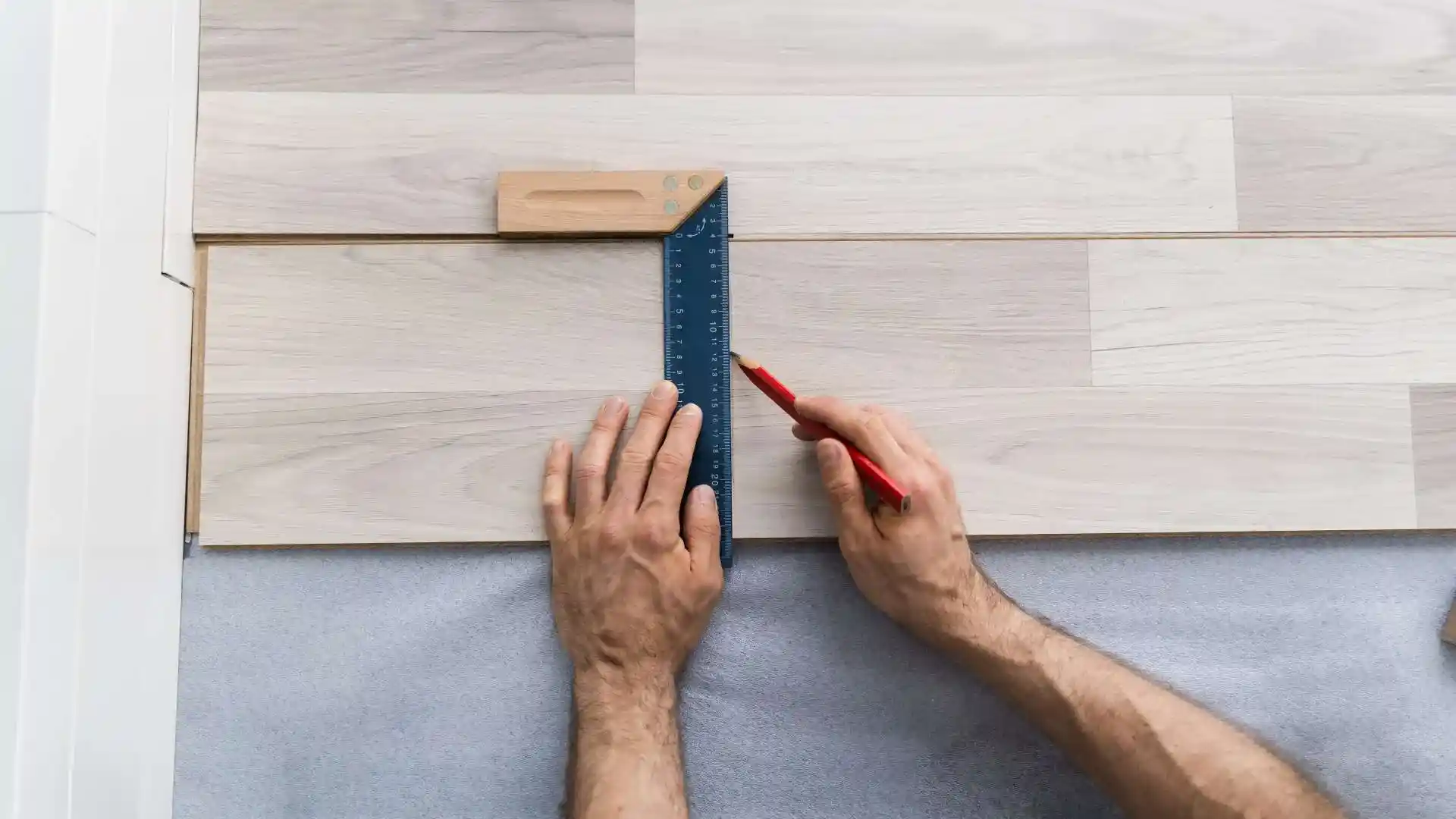 A person uses a square ruler and red pencil to mark a straight line on light-colored wood flooring, preparing for precise interior trim installation.