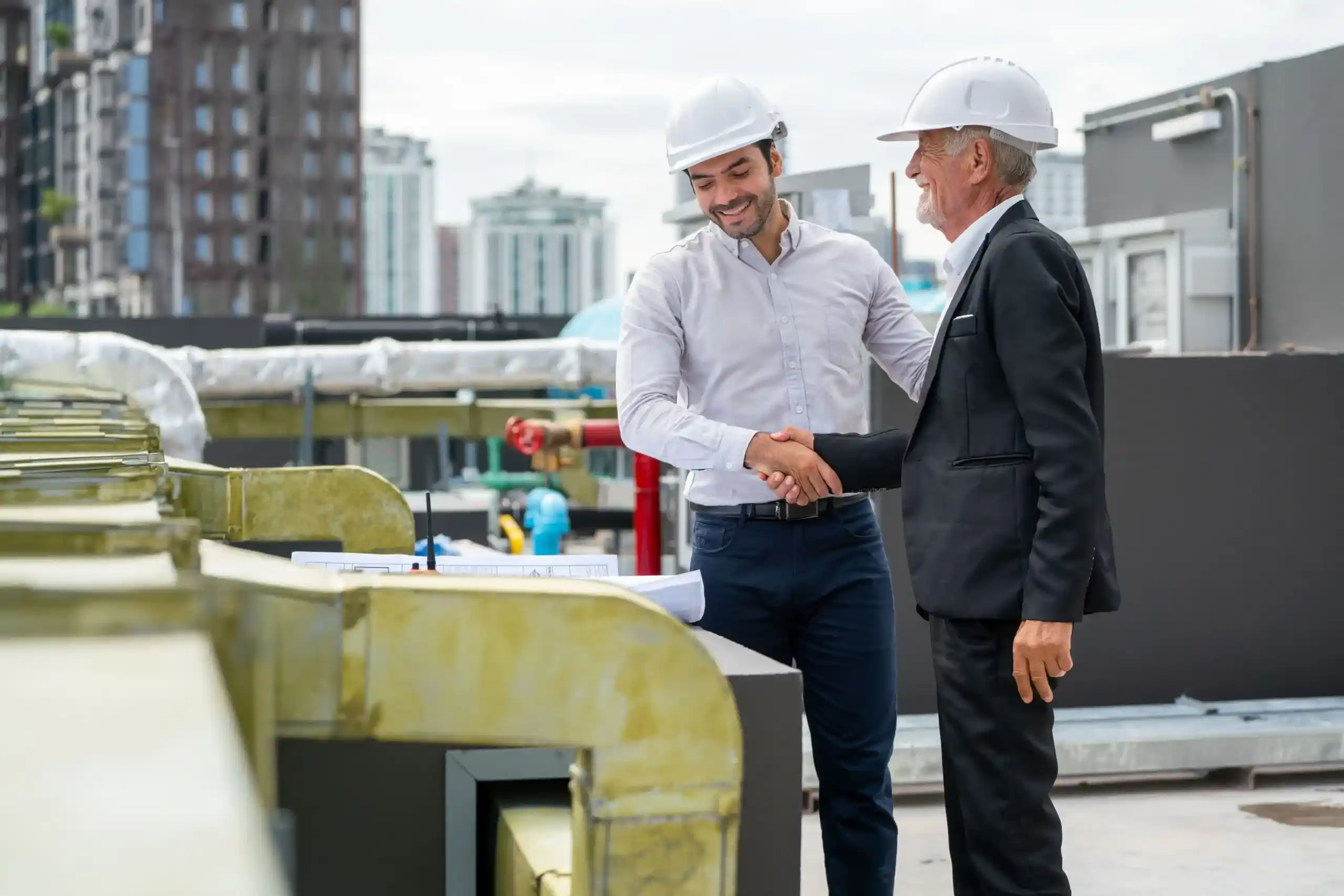 Two men wearing hard hats and business attire shake hands at a construction site, with city buildings in the background—highlighting the importance of securing construction permits in Canada.