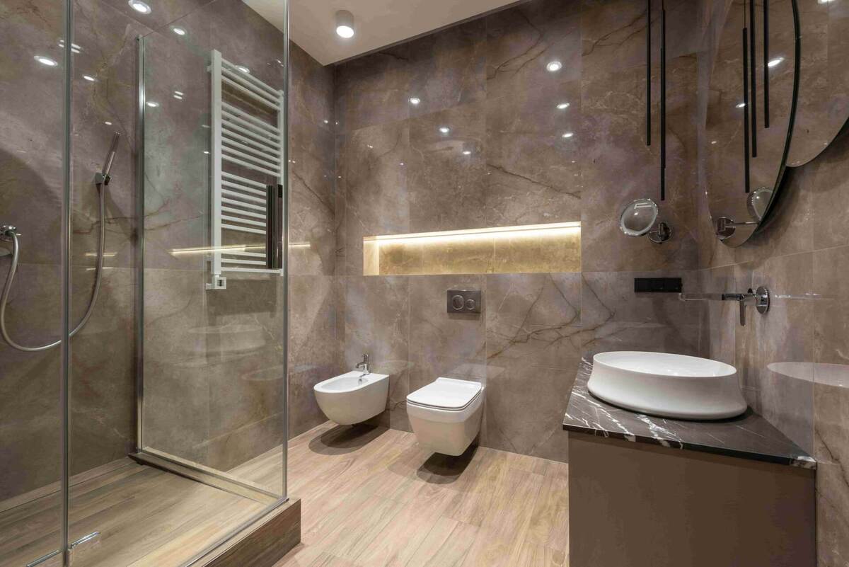 Modern bathroom with marble walls, wood-look floor, glass shower, wall-mounted toilet and bidet, vessel sink on a vanity, large mirrors, and illuminated shelf—perfect inspiration if you’re wondering How Long Does a Bathroom Renovation Take in New Westminster?.