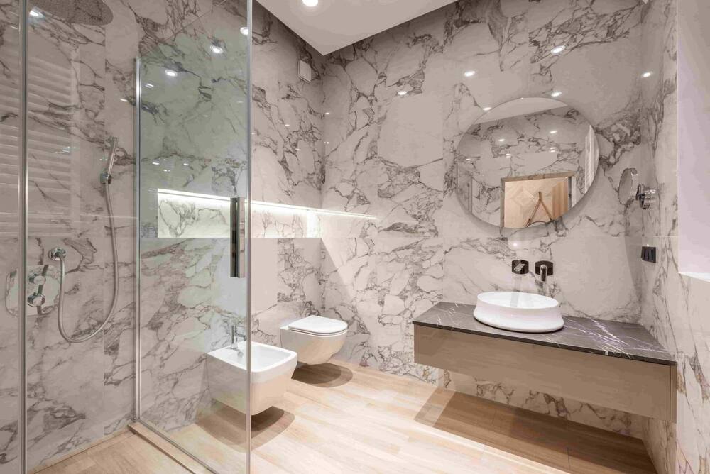 Modern bathroom with marble walls—one of the best materials to use for a bathroom renovation in a Richmond home—plus a glass shower, wall-mounted toilet and bidet, round mirror, floating vanity with vessel sink, and wood-look flooring.