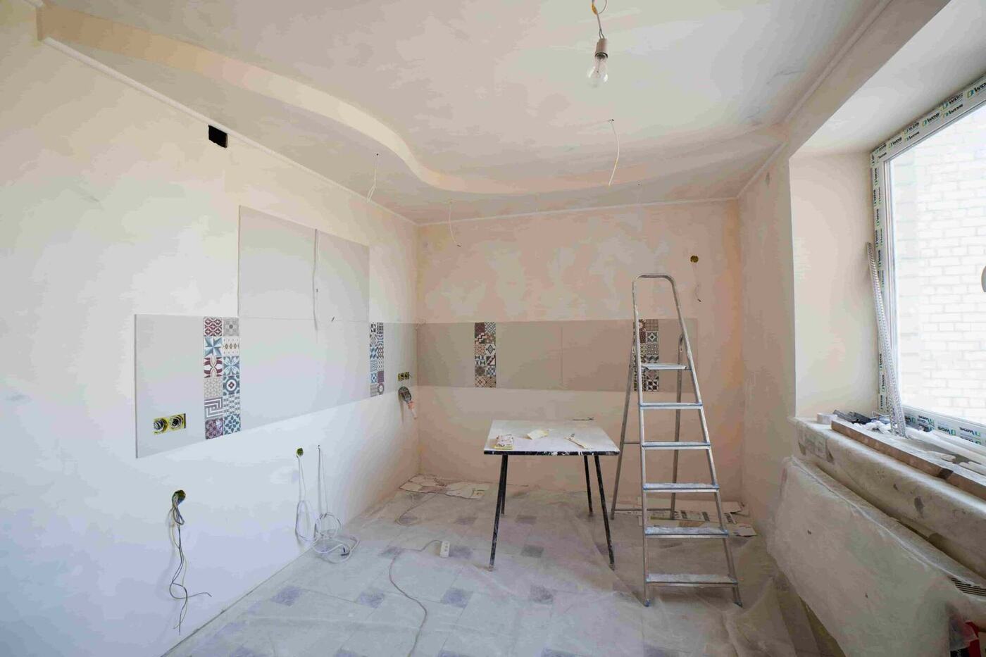 A room under renovation with unfinished walls, a step ladder, a table with tools, exposed wiring, and a large window letting in natural light—perfect inspiration for budget-friendly home renovation ideas for Coquitlam homes.