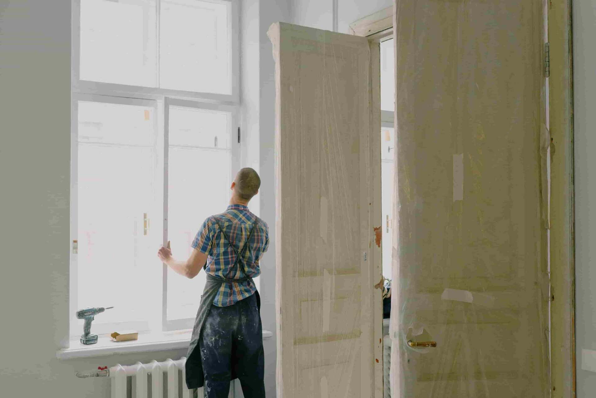A person in work clothes installs or repairs a window in a room with covered doors and construction tools nearby, showcasing a Step-by-Step Guide to Renovating Older Multifamily Properties.