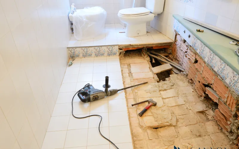 Bathroom under renovation with tiles and floor partially removed; contractors for bathroom renovations have left tools including a drill and hammer on the floor near exposed pipes and bricks.