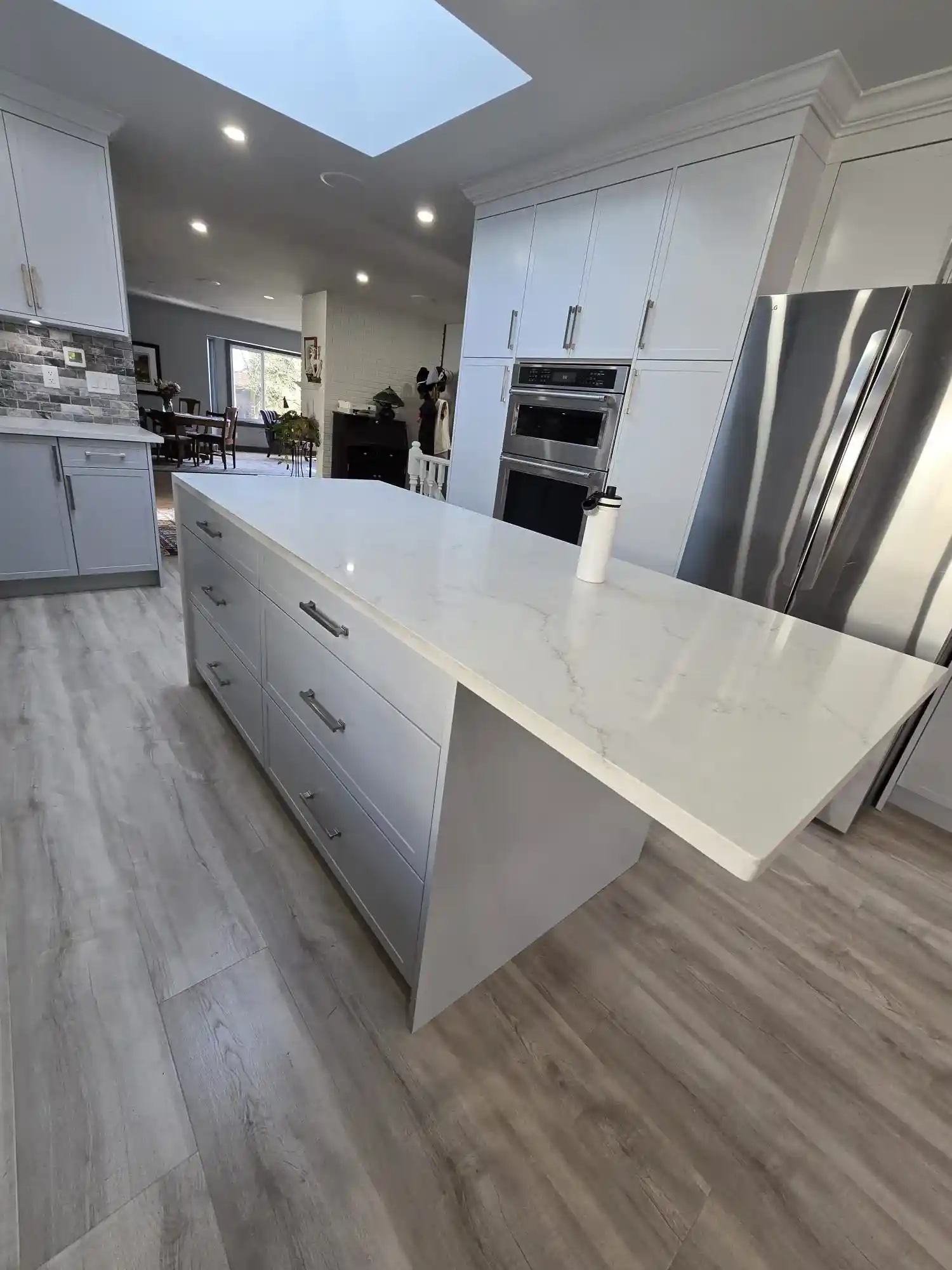 Modern kitchen with a large white marble island, stainless steel appliances, light wood flooring, and white cabinetry—perfect for custom home builds. A water bottle sits on the island countertop.