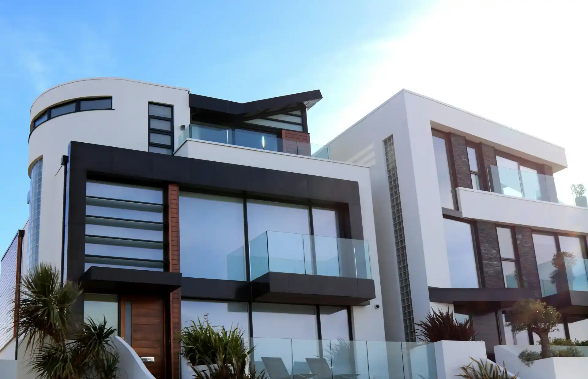 Modern white and black multi-story house with large glass balconies, flat roofs, and palm landscaping—showcasing a recent complete building envelope restoration—under a clear blue sky.