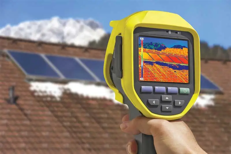 A person holds a thermal imaging camera aimed at roof solar panels, displaying a thermal view of heat distribution on the camera screen—an essential tool for detecting issues that could indicate the need for roof repair.