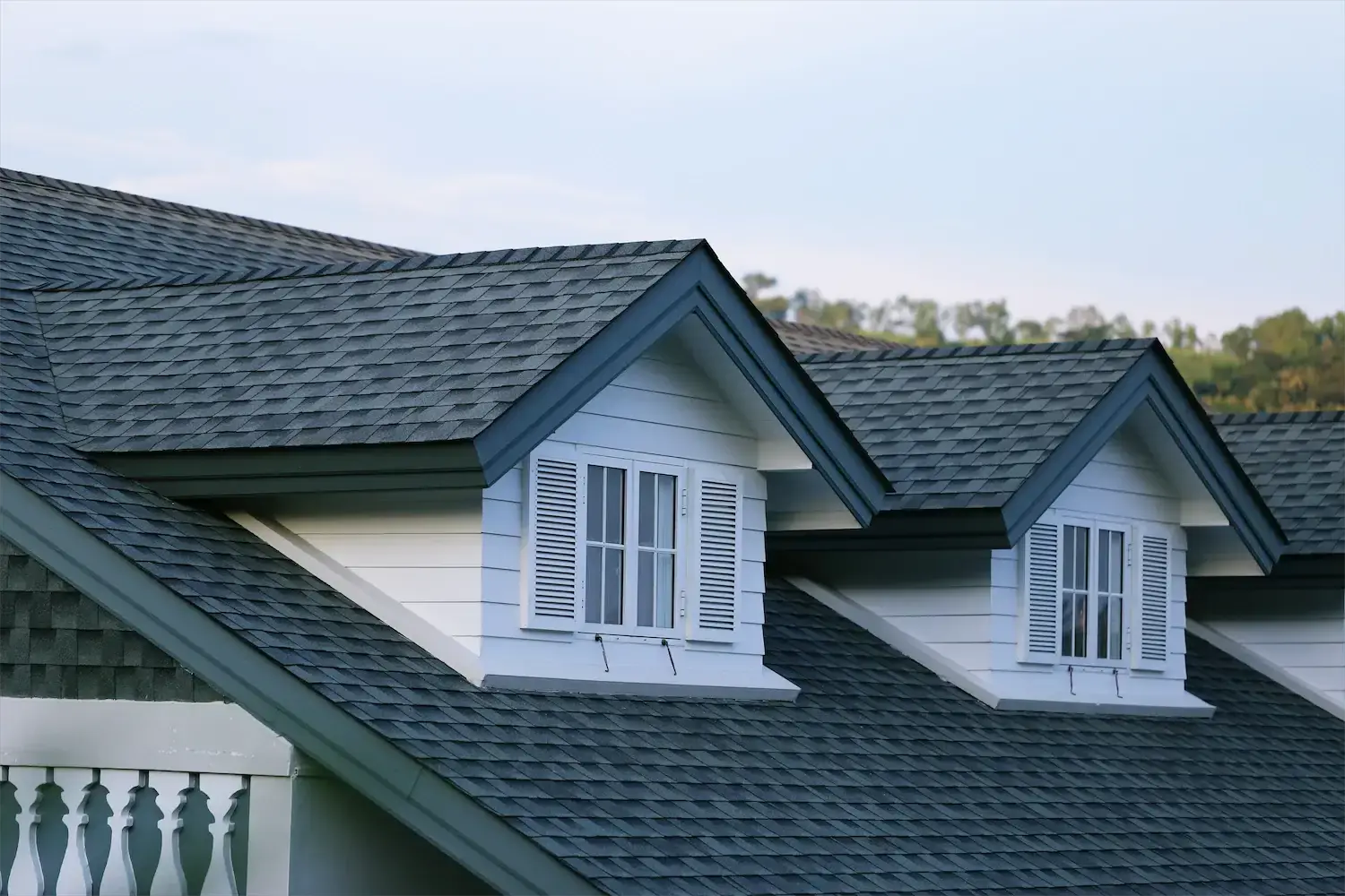 Close-up view of two dormer windows with white shutters on a gray shingled roof, showcasing the fresh look after a recent roof replacement, with trees and sky in the background.