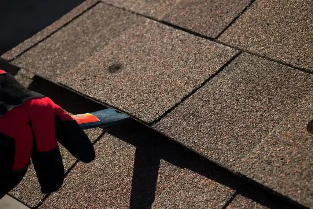 A gloved hand lifts an asphalt roof shingle with a pry bar, exposing the edge of the shingle underneath in sunlight—a careful step in effective roof repair.