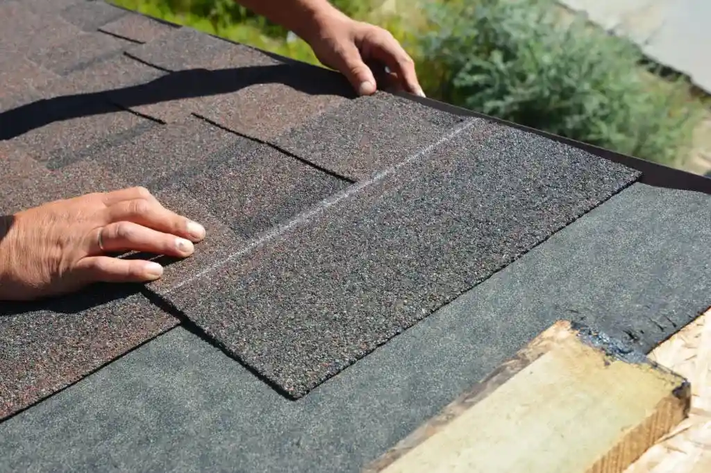 Hands installing asphalt shingles on a roof during a roof replacement, with underlayment visible and greenery in the background.
