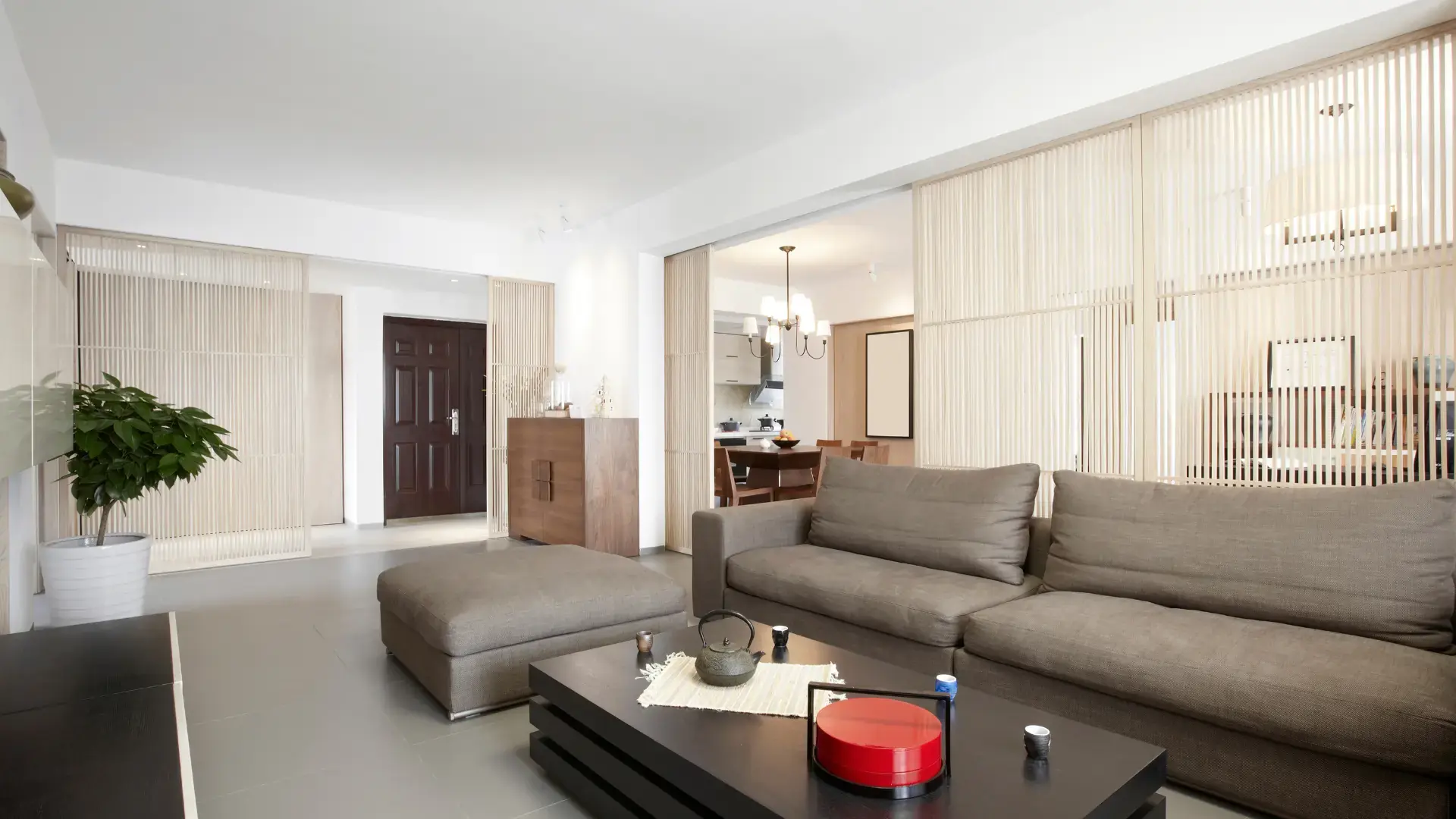 Modern living room with a gray sofa, matching ottoman, and black coffee table. Sliding wooden panels separate the dining area. A potted plant stands near a dark wooden door in the background, reflecting exquisite style seen in home renovations across Surrey.