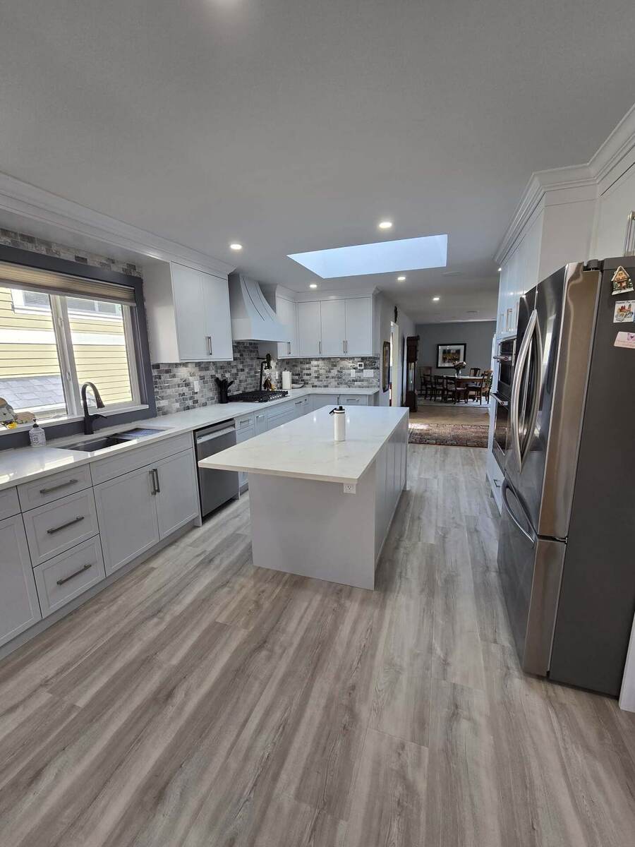 Modern kitchen with white cabinets, a large island, stainless steel appliances, gray backsplash, and wood-look flooring under a skylight—meticulously cared for with regular property maintenance.