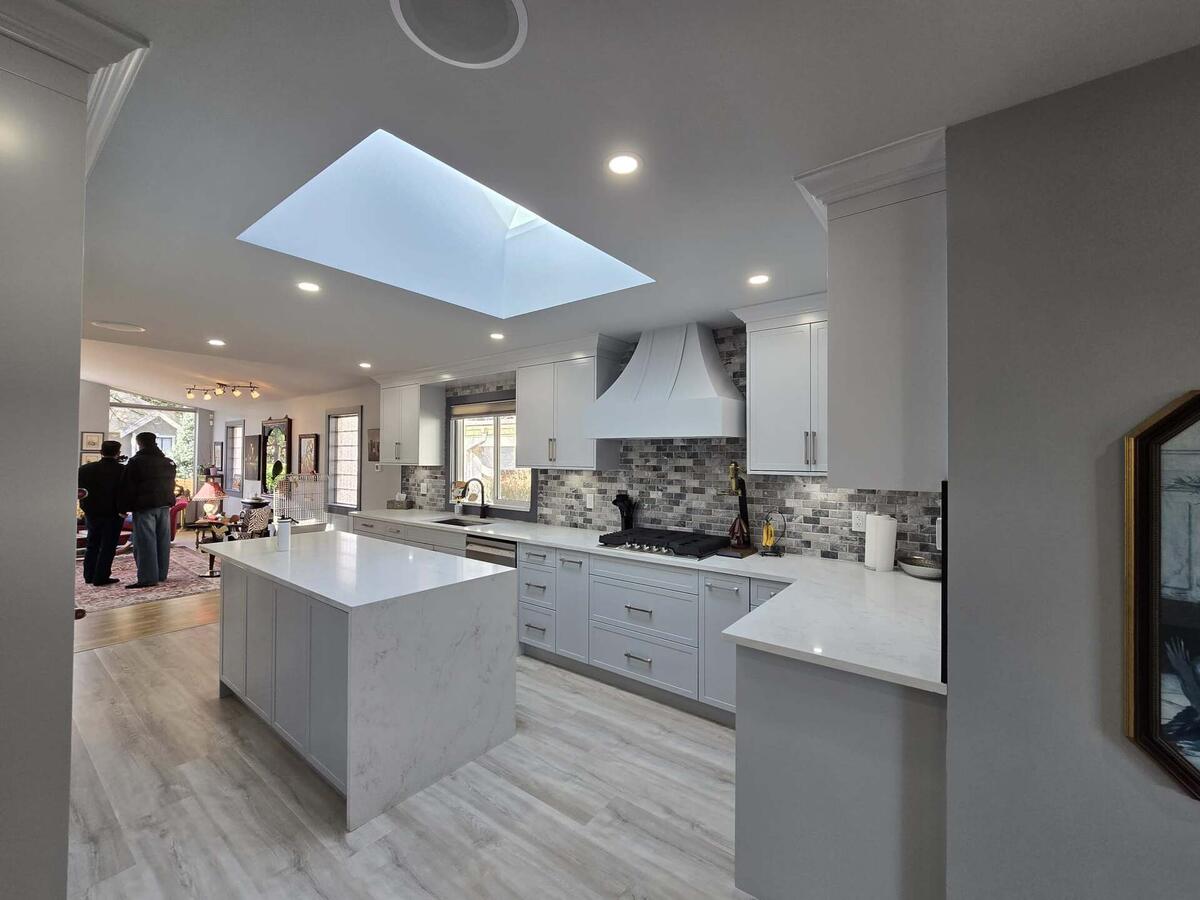 Modern kitchen with white cabinets, marble island, and a skylight; two people are seen standing in the adjacent living area, highlighting the benefits of attentive property maintenance.