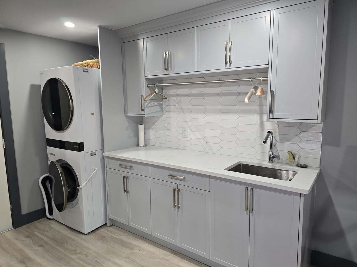 Modern laundry room with stacked washer and dryer, light gray cabinets, white hexagon tile backsplash, and a stainless steel sink—ideal for easy property maintenance.