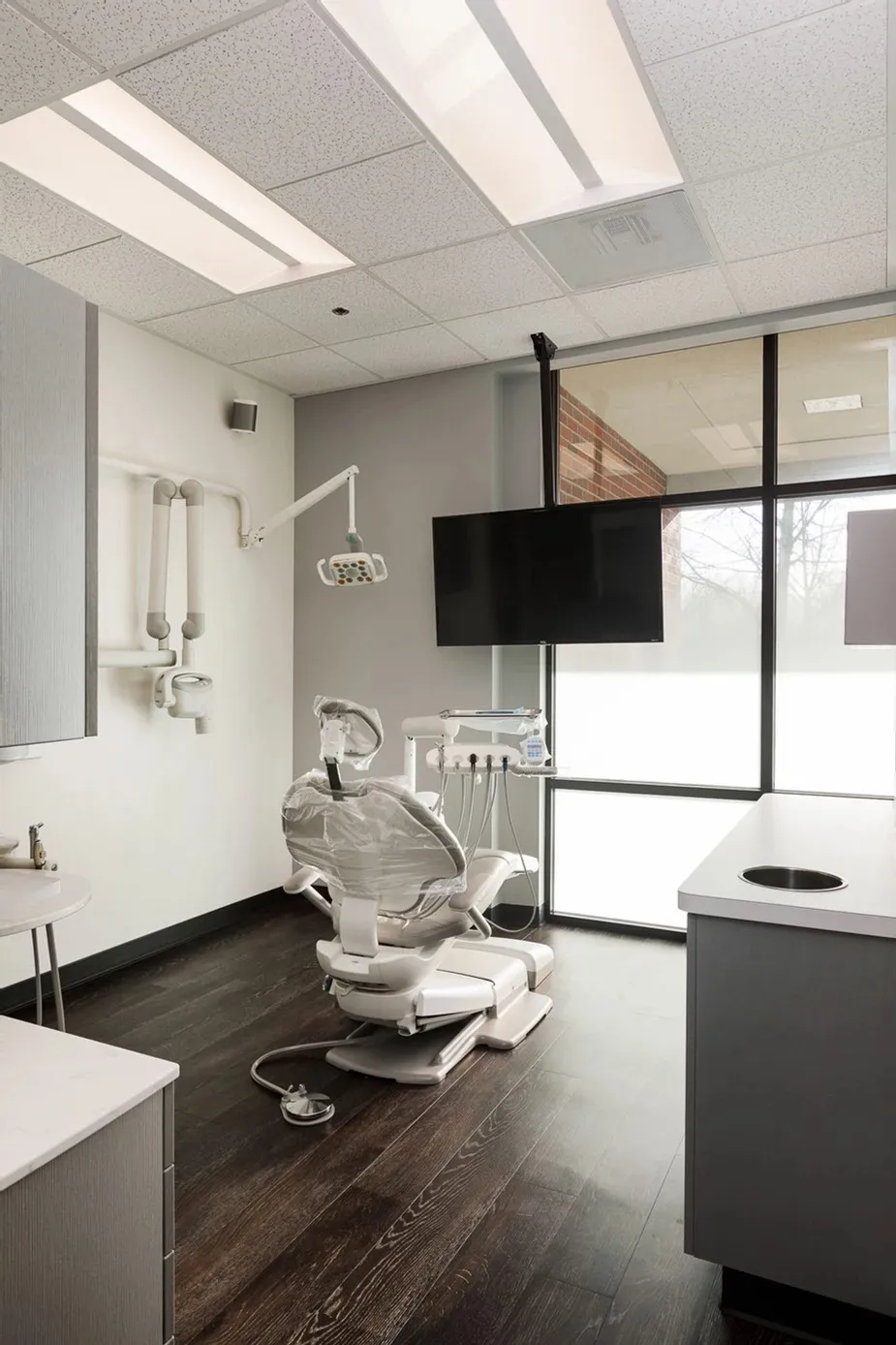 A modern dental exam room, recently updated through clinic renovations Canada, features a dental chair, overhead light, equipment, large monitor, and a floor-to-ceiling window bringing in natural light.