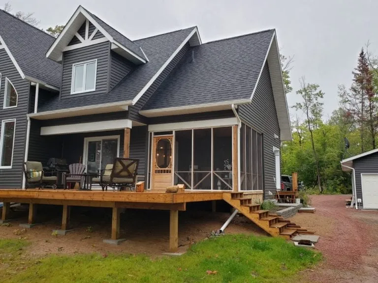A two-story gray house with a covered porch, wooden deck, outdoor chairs, and screened-in area boasts professional siding/deck services, all set in a wooded area with a gravel driveway.