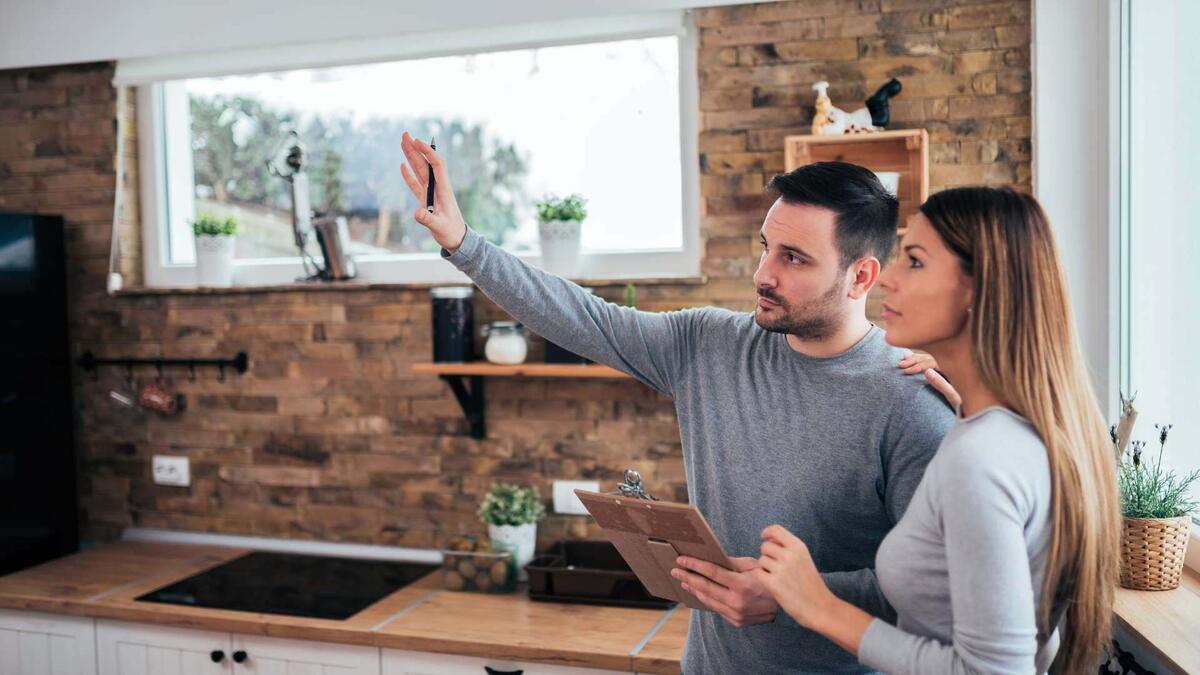 A man and woman stand in a kitchen with a brick wall; the man is gesturing upward while holding a clipboard.