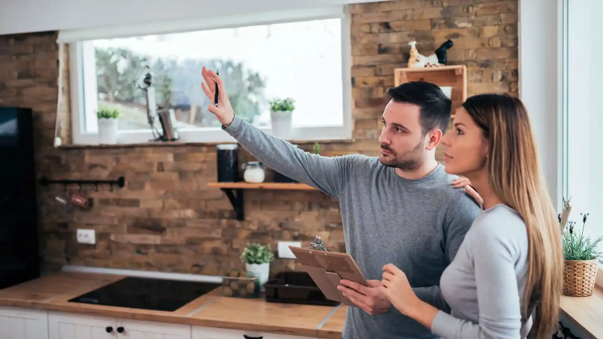 A man holding a clipboard gestures while discussing property maintenance with a woman in a modern kitchen featuring brick walls and wooden shelves.